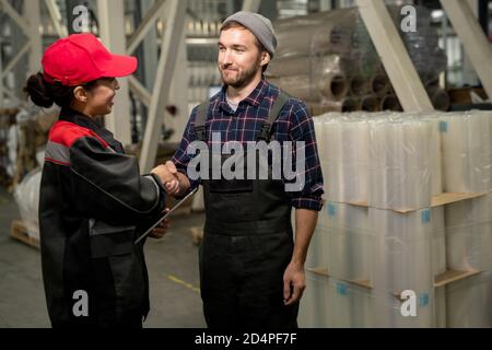 Happy young male engineer in overalls and checkered shirt shaking colleague hand Stock Photo