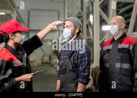 Young female medical worker in protective mask measuring temperature of workers Stock Photo
