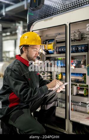 Contemporary young engineer of factory in workwear and hardhat using tablet Stock Photo