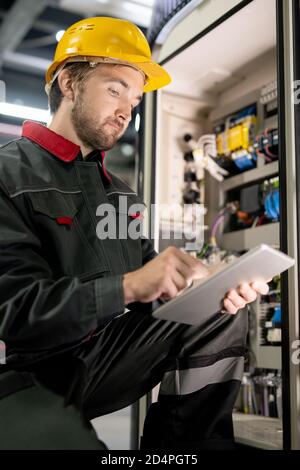 Young bearded engineer in workwear and hardhat checking settings of machine Stock Photo