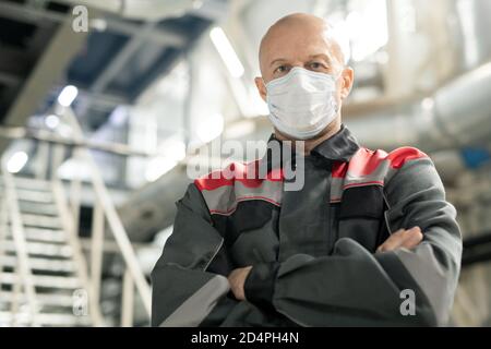 Bald mature worker in uniform and protective mask standing against staircase Stock Photo