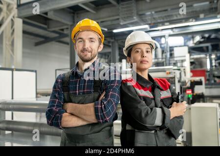 Two young confident workers in helmets and workwear standing in large workshop Stock Photo