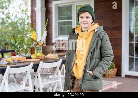 Cute schoolboy in warm casualwear looking at you against served table and chairs Stock Photo