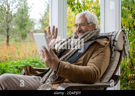 Happy senior man with white hair waving hand while sitting in rocking chair Stock Photo