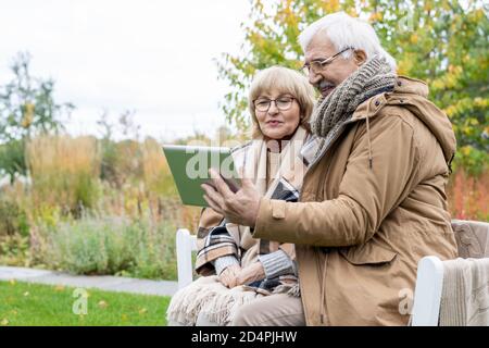 Elderly husband and wife with digital tablet watching online movie on bench Stock Photo