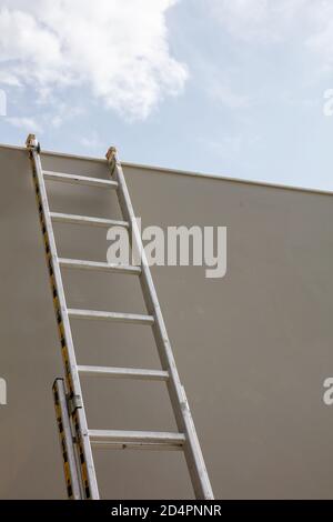 Maintenance concept. Ladder aluminium, light, silver color put on grey wall. Support for repairing with safety. Low angle view, vertical, blue sky bac Stock Photo