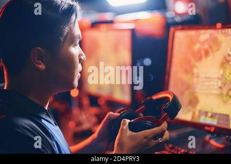 Side view of a focused asian guy, male professional cybersport gamer holding headphones and looking at PC screen while playing online video game. Participating in eSport tournament Stock Photo