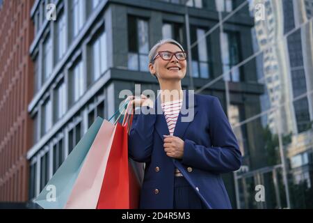 Happy mature woman holding shopping bags. Portrait of confident asian businesswoman wearing stylish eyeglasses walking on the street Stock Photo