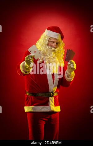 Santa Claus holds playing cards in his hands and poses on a dark red background Stock Photo