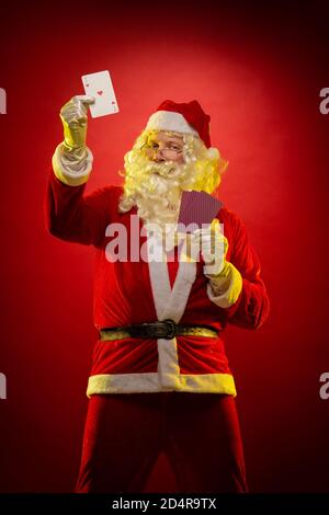 Santa Claus holds playing cards in his hands and poses on a dark red background Stock Photo
