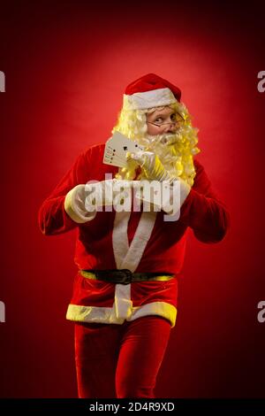 Santa Claus holds playing cards in his hands and poses on a dark red background Stock Photo