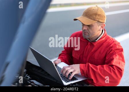 Handsome auto mechanic or road assistance worker in uniform repairing engine of the broken car on the road Stock Photo