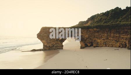 Sunrise of green cliff at ocean bay sand coast aerial view. Mesmerizing sun rize at Batu Bolong rock wall with giant hole at Bawana Beach, Sumba Island, Indonesia. Cinematic soft light drone shot Stock Photo