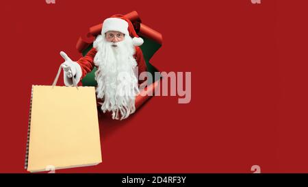 Santa man holds shops packet in hand through a paper hole. Bearded man in santa hat looking through hole on red paper. Stock Photo