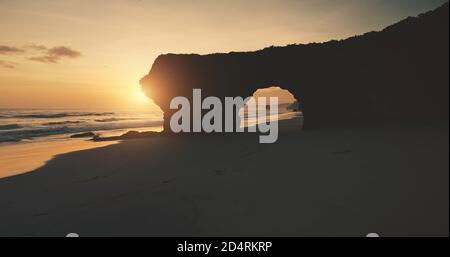 Sunlight silhouette of hole at rocky wall on sand ocean coast. Aerial view of Bawana Beach tourist attraction. Unique geological formation from cliff. Indonesia landmark soft sun set light Stock Photo