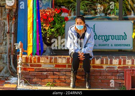 Ellicott City, MD, USA 10/07/2020: A young hispanic woman working at a jewelery shop is sitting on window sill during her break. She is wearing face m Stock Photo