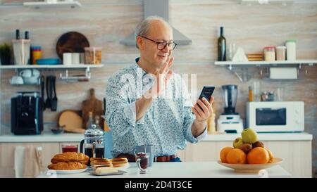 Senior man shacking hand during a video call in kitchen using smartphone. Elderly person using internet online chat technology video webcam making a video call connection camera communication conference call Stock Photo