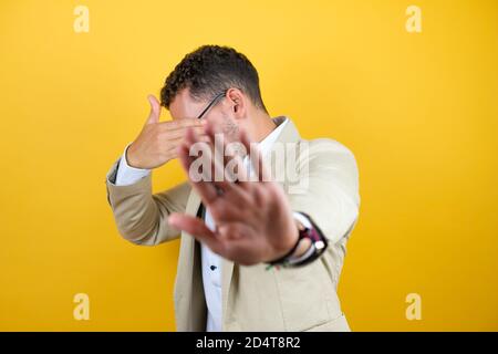 Young handsome businessman wearing suit over isolated yellow background covering eyes with hands and doing stop gesture with sad and fear expression. Stock Photo