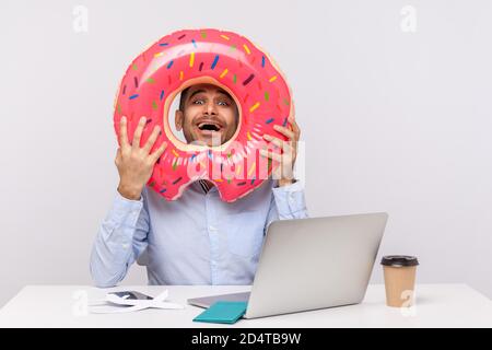 Extremely happy man looking through rubber ring and smiling excitedly, sitting in office workplace with passport on desk, celebrating planned travel v Stock Photo