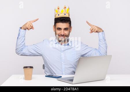 Look, I'm king! Proud authoritative businessman pointing at crown on head while sitting in office workplace, feeling self-confident and ambitious to c Stock Photo