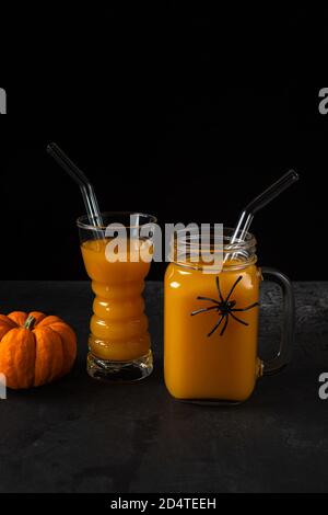 Non-alcoholic pumpkin mocktail in drinking glass and square jar with glass straws and whole orange pumpkin on black textured surface. Halloween spider Stock Photo