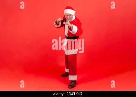Full length portrait of happy purposeful gray bearded man in red santa claus costume and eyeglasses holding clenched fists up ready to boxing. Indoor Stock Photo