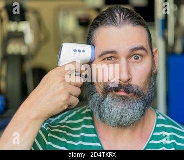 Bearded man with long dark hairs points an infrared thermometer at his face as if it's a gun. Self care, medical and funny concept.  Stock Photo