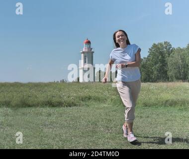 happy caucasian woman running to a viewer. green meadow and old white lighthouse on the background. local travel and weekend staycations. calm and peaceful place. Stock Photo