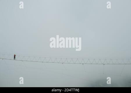 Suspencion bridge high up in cloudy mountains and man standing on it Stock Photo