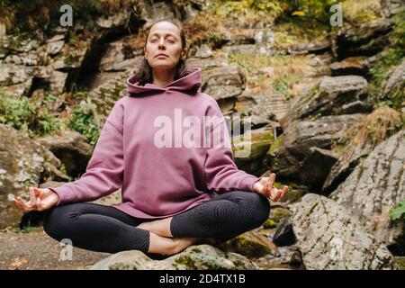 Peaceful young woman sitting on big stone cross-legged, meditating Stock Photo