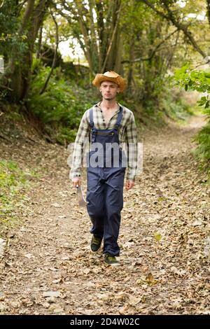 Blond-haired man with a billhook, wearing dungarees and a straw hat. Some movement blur on man. Stock Photo