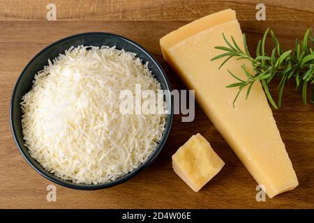 Grated Parmesan or Grana Padano in a ceramic bowl, wedge of hard cheese and rosemary on a brown cutting board. Delicious ingredient for pizza, sandwic Stock Photo