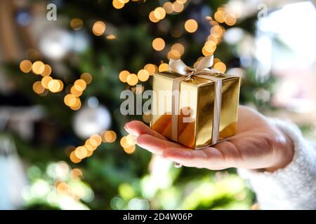 Close up of hand holding a golden gift box with Christmas tree and lights on the background, present and Christmas holiday concept Stock Photo
