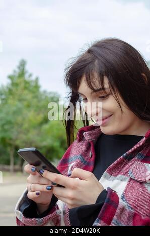 Portrait of a young woman writing a message on the phone. Woman with long flowing hair in a plaid shirt outdoors. Lifestyle Stock Photo