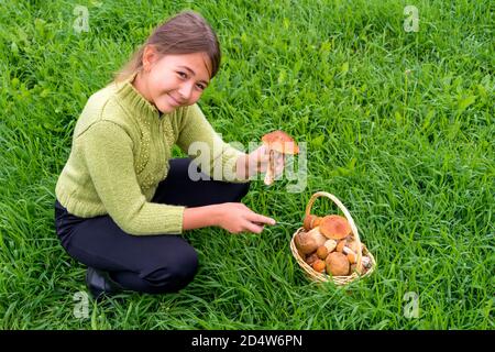 Picking mushrooms. Caucasian girl in green knitted sweater cut fresh edible young mushroom boletus edulis hunting knife near wicker basket with edible mushrooms on a forest lawn, close up. Red cap boletus, mushroom hunting. Space for text.harvest Stock Photo