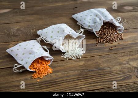 Three types of cereals buckwheat, rice and lentils in canvas bags. They lie on a wooden background. Grains are scattered on the table. Organic healthy Stock Photo