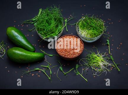 Microgreens: green peas and onion sprouts, flax seeds in glass bowls and avocado on black background. Healthy vegetarian food and diet concept. Stock Photo