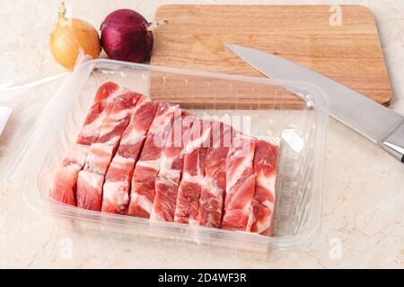 Fresh raw pork belly pieces in a disposable transparent plastic food container near wood cutting board and sharp knife on kitchen table. Cook at home. Stock Photo
