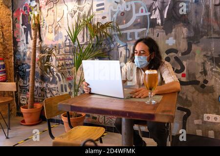 Man using a laptop inside a modern bar. He is wearing a medical mask. Space for text. Stock Photo