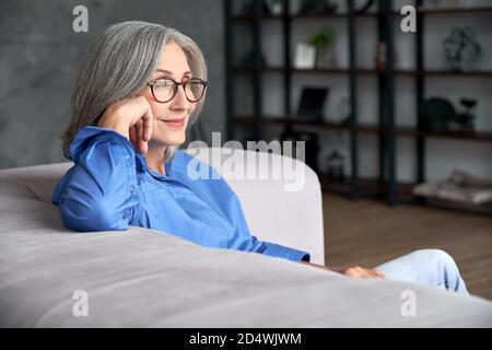 Beautiful mature older woman wearing glasses relaxing sitting on couch at home. Stock Photo