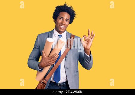 Handsome african american man with afro hair holding paper blueprints doing ok sign with fingers, smiling friendly gesturing excellent symbol Stock Photo