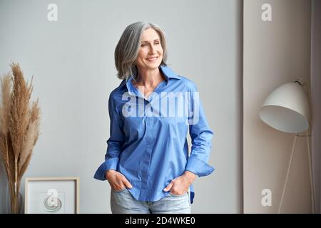Smiling confident mature older woman standing indoors looking at window. Stock Photo