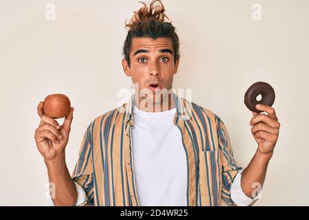 Young hispanic man holding red apple and donut in shock face, looking skeptical and sarcastic, surprised with open mouth Stock Photo