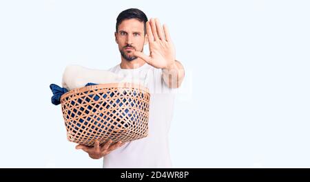 Young handsome man holding laundry basket with open hand doing stop sign with serious and confident expression, defense gesture Stock Photo