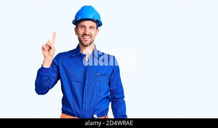 Young handsome man wearing worker uniform and hardhat pointing finger up with successful idea. exited and happy. number one. Stock Photo