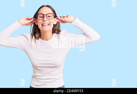 Beautiful brunette young woman wearing casual white sweater and glasses smiling pulling ears with fingers, funny gesture. audition problem Stock Photo