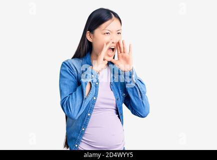 Young beautiful chinese woman pregnant expecting baby shouting angry out loud with hands over mouth Stock Photo