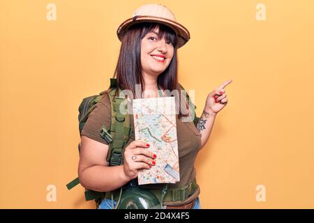 Young plus size woman wearing explorer hat holding map smiling happy pointing with hand and finger to the side Stock Photo