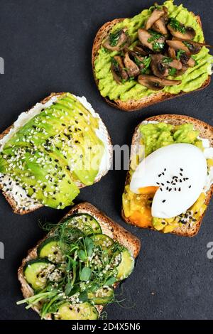 Assortment of avocado toasts. Healthy toast with avocado, egg, fried mushrooms, cucumber and microgreens garnished with sesame seeds. Top view on blac Stock Photo