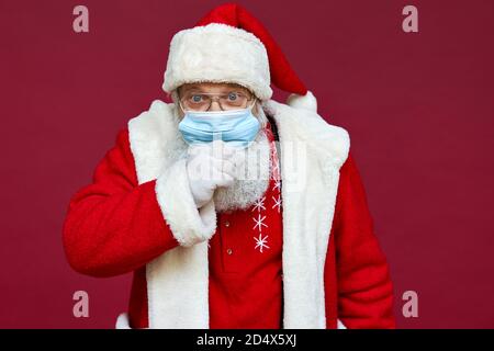 Sick ill Santa Claus wearing face mask coughing isolated on red background. Stock Photo